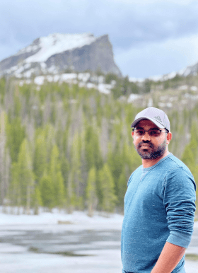 Dinesh is wearing a grey hat, blue shirt, and sunglasses, and is standing in front of a landscape with a forest and mountain in the background.